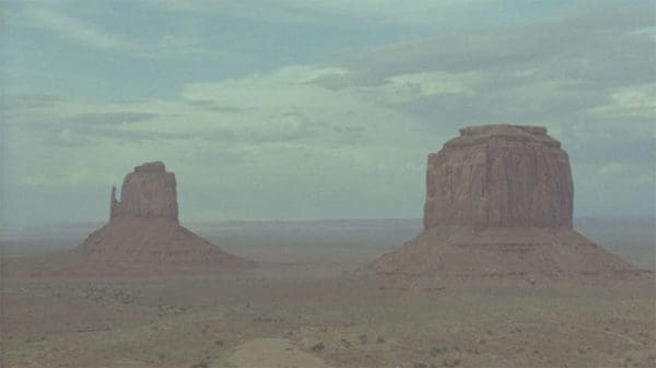 Two large, flat-topped rock formations rise from a vast, arid desert landscape under a cloudy sky.