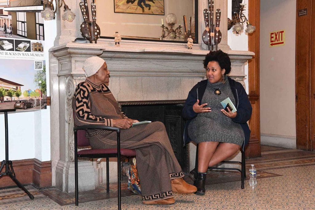 Two women talk in chairs in front of a fireplace