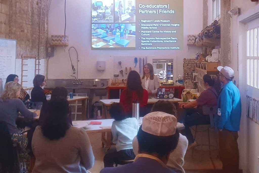 a woman speaks to a group seated at work tables, looking at a presentation on "Co-educators, Partners, and Friends."