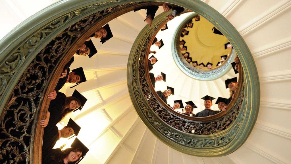 Looking up the spiral staircase at the Peabody Institute, where students in graduation attire line the steps.