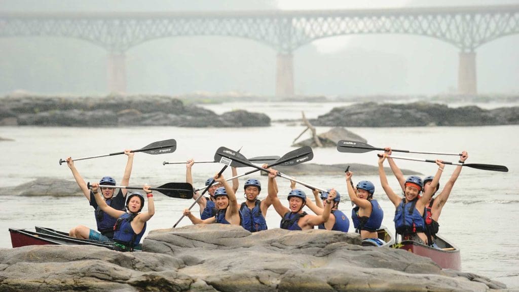 On a pre-orientation outdoor trip in Harper's Ferry, students gather in kayaks on the water, raising their paddles overhead with a bridge in the background.