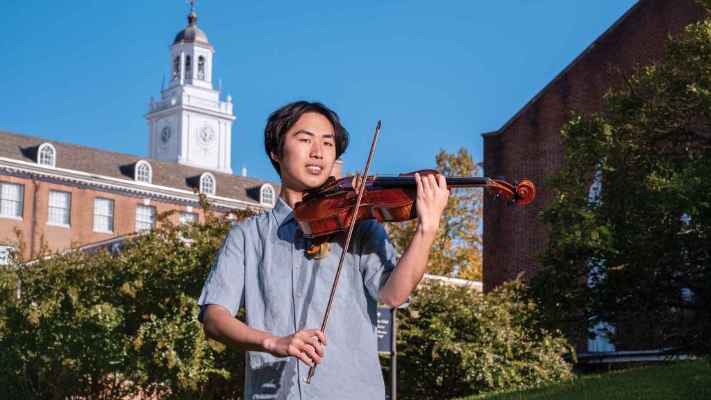 Henry Hung holds holds viola getting ready to play outside on Homewood campus with Gilman Hall tower in the background.