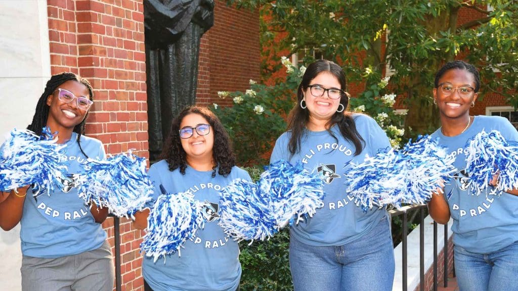 four smiling students in FLI/JHU t-shirts, pom-poms in hand, cheerfully welcoming new FLI students with