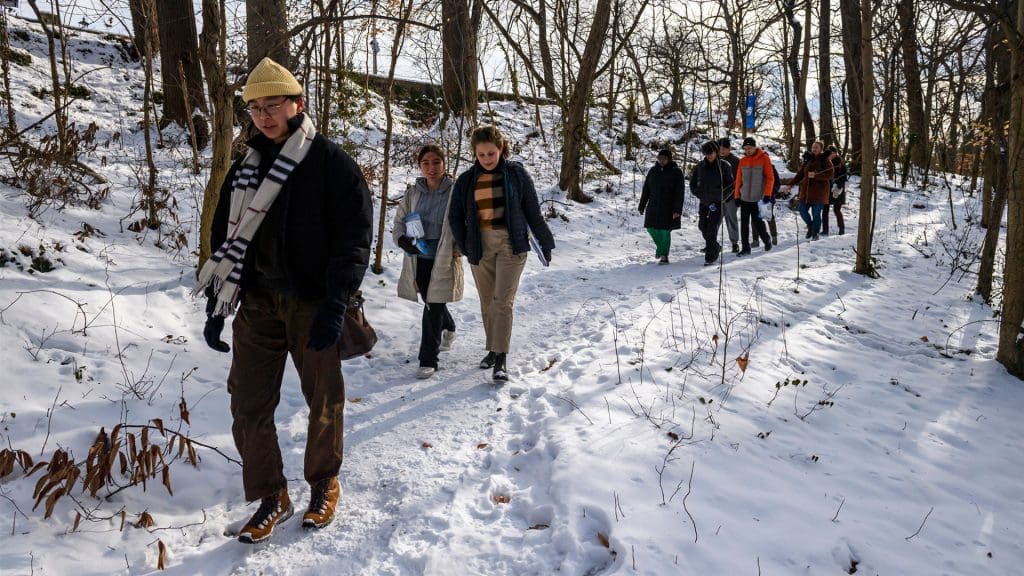 Teacher (a doctural student) leads multiple students through snowy woods, all are dressed in winter jackets.