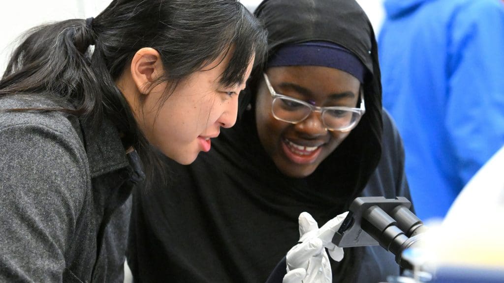 Teacher and student discuss while both look over a microscope.