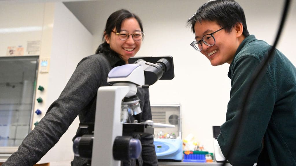 The two teachers smile while looking at a microscope in a lab.