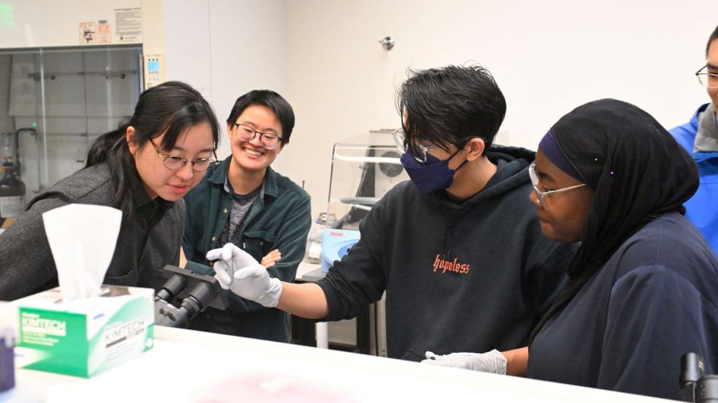 Teachers and students discuss while looking over a microscope.