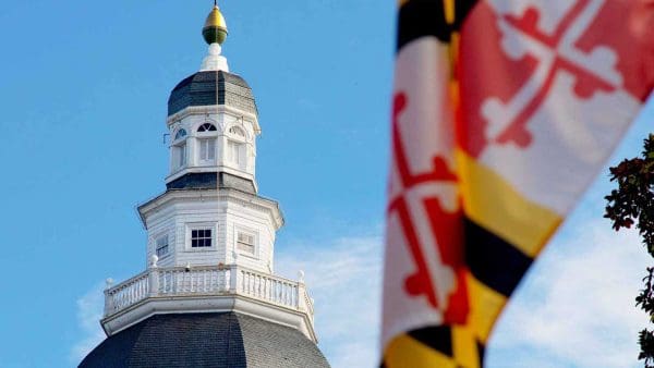 The Maryland flag in the foreground and the Maryland State House in Annapolis in the background on a sunny day.