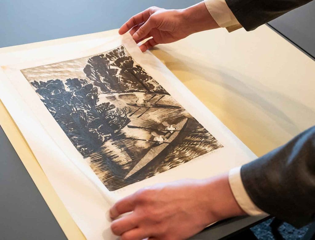 close-up of hands lifting a black, gray, and white lithograph print of two kayaks, each with two people, going down a stream with trees on either side
