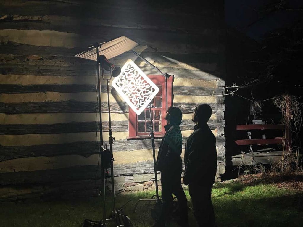 the shapes of two people in the dark, standing under a light next to a wood cabin