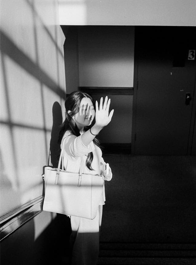 Black and white photo of a young woman raising her hand to block sunlight streaming through a window, its frame casting shadows across her.