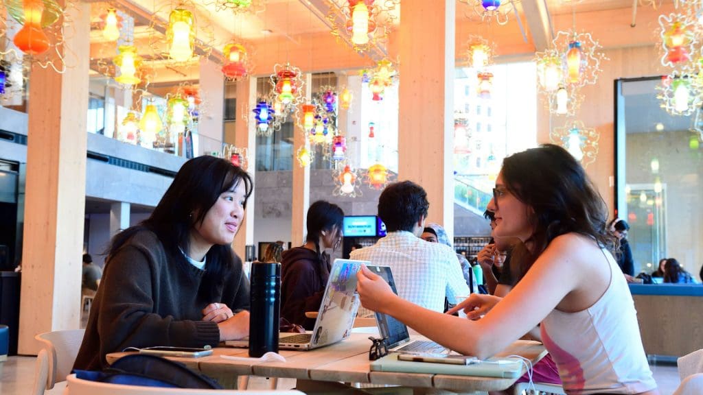 Two students in the main seating area of The Bloomberg Student Center talking to eachother while studying.