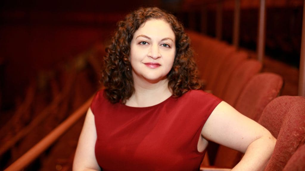 Jill Rafson in a red shirt, red theater chairs are behind her.
