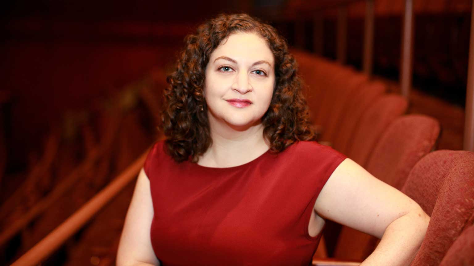 Jill Rafson in a red shirt, red theater chairs are behind her.