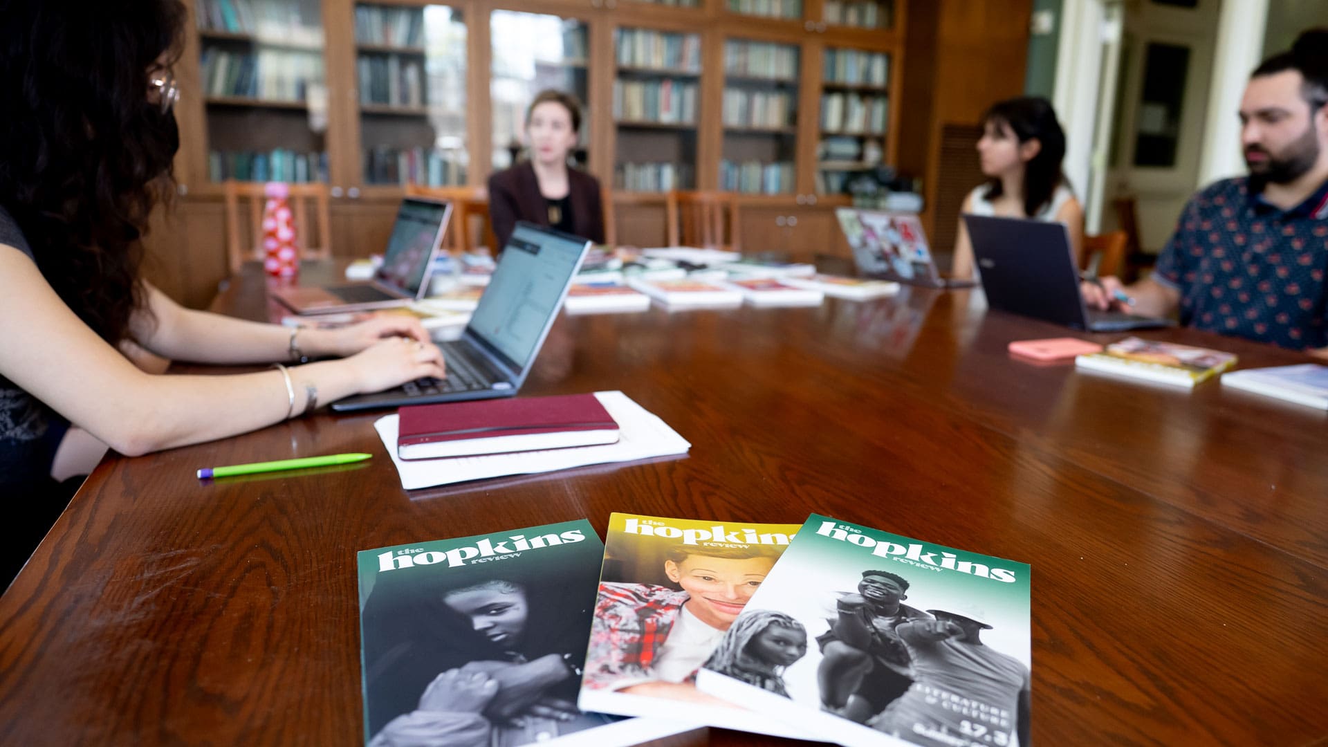Hopkins Review meeting held on big wooden table, student workers and Dora Malech are in the background out of focus. In the foreground is 3 issues of the Hopkins Review.